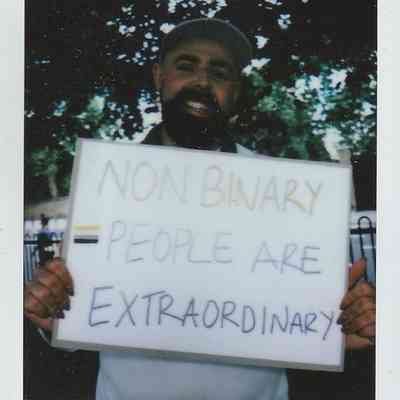 A person holds a sign saying "Non-Binary People Are Extraordinary" with trees in the background.