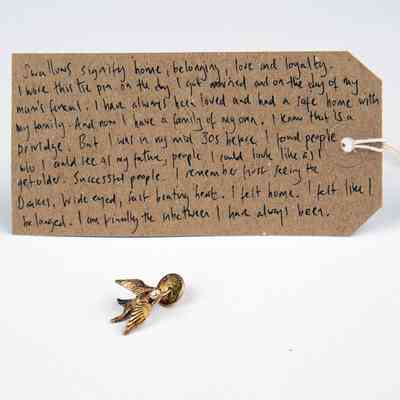 A small golden pin which is a swallow. Behind it is a brown luggae tag with writing on it.
