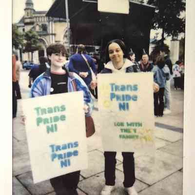 Two people stand holding Trans Pride NI signs, with a historic building in the background.