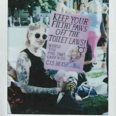 A person sat on the grass holding a placard which says "Keep Your Filthy Paws Off The Toilet Laws".