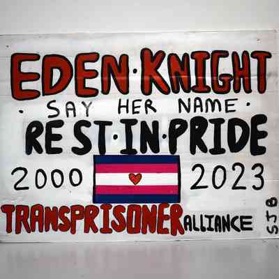 A white plastic placard reading "EDEN KNIGHT SAY HER NAME REST IN PRIDE 2000 - 2023 TRANSPRISONER ALLIANCE SJB" in red and black, handwritten text. Between the dates, there is a trans flag with a heart in the centre.