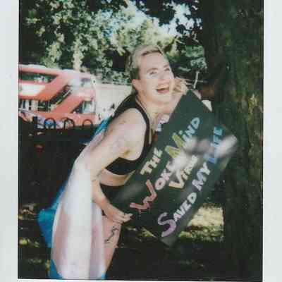 A smiling person stands under a tree, holding a colorful sign with text, next to a red bus.