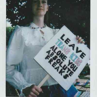 A person holding a sign advocating for trans kids under a tree in London setting.