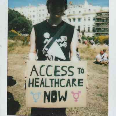 A person holds a sign reading "ACCESS TO HEALTHCARE NOW" during a sunny outdoor protest.