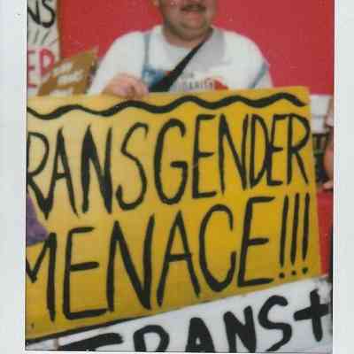 A person with glasses smiles while holding a yellow sign with bold text reading, "TRANSGENDER MENACE".