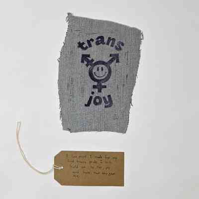 A lino cut print on a scrap of blue denim. Text "trans joy" around the trans logo (stylised with a smiley face in the centre). With a brown handwritten luggage tag.