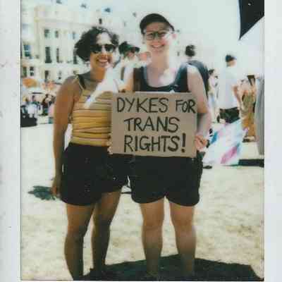 Two individuals stand together holding a sign which says "DYKES FOR TRANS RIGHTS".