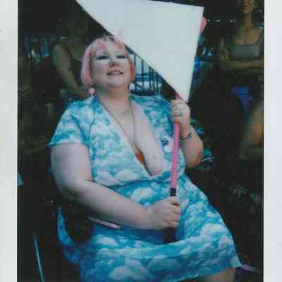 A person with pink hair smiles while holding a large triangular sign, surrounded by others.