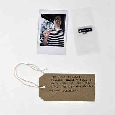 A Polaroid of a person in a striped top wearing glasses and holding a small badge, a blue enamel 'SHE/THEY' pin badge and a handwritten brown luggage tag.
