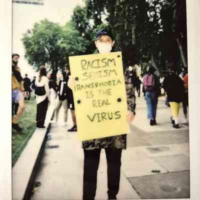 A person holds a yellow sign reading "Racism, Sexism, Transphobia Is the Real Virus".
