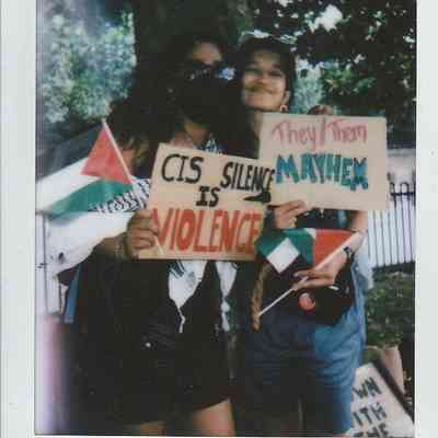 Two people hold protest signs, one reads "Cis Silence is Violence," and the other says "They/Them Mayhem" under a tree.