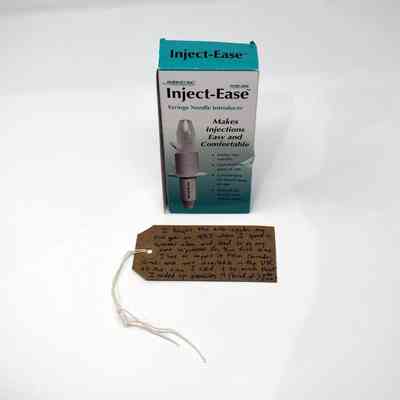 A box of Inject-Ease which is turquoise and white with a photograph of the syringe needle introducer on it. Underneath the box is a brown tag with writing on it.