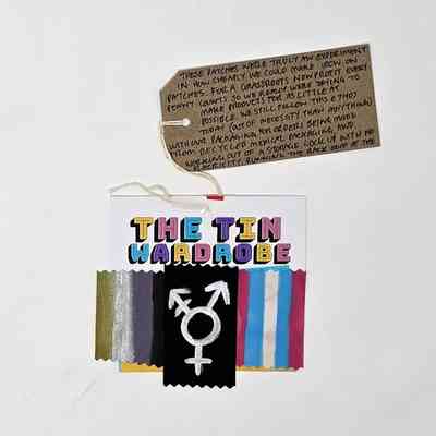The fabric patches depicting the Non-Binary Flag, Trans Flag and the Trans Symbol, mounted on a card titled "THE TIN WARDROBE", with a brown handwritten luggage tag.