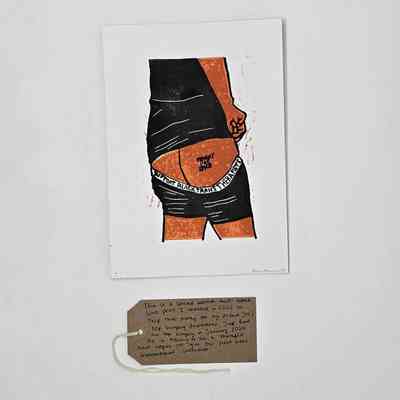 A print of a River Manning illustration. It shows the rear torso and upper legs of a person wearing a black top and shorts. Their shorts are partially pulled down to reveal at a tattoo "TRANNY FAG DYKE" on their right buttock. With a brown, handwritten luggage tag.