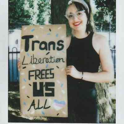 A person stands by a tree, smiling, holding a sign saying, "Trans Liberation Frees Us All".
