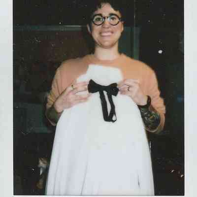 A smiling person stands holding a white garment with a black bow in their hands inside a room.