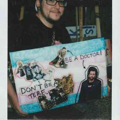 A person wearing a cap and glasses holds a colorful collage poster with various images and text.