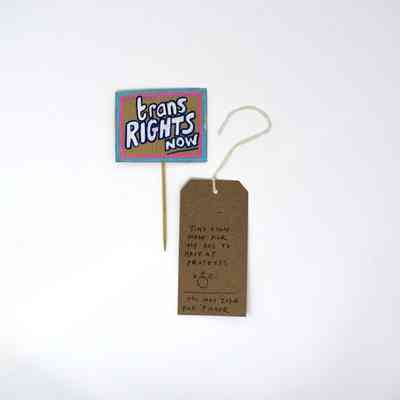 The image displays a small protest sign reading "Trans Rights Now" with a brown tag.