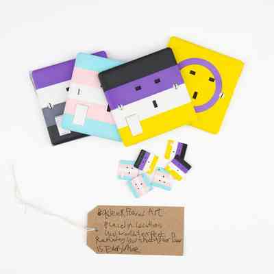 Several 3D printed 3-pin socket facias coloured with the Asexual, Trans, Non-Binary and Intersex flag colours. With a brown, handwritten luggage tag.