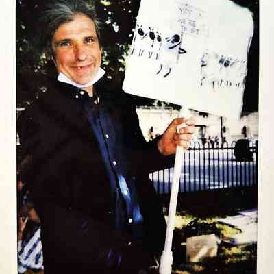 A man stands outdoors holding a protest sign with sheep illustrations, smiling confidently at the camera.