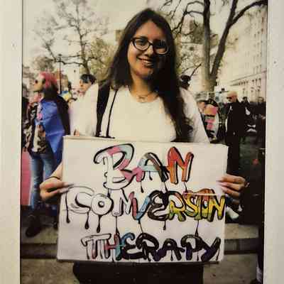 A person holds a colorful sign reading "Ban Conversion Therapy" in a busy outdoor setting.