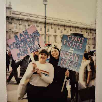 People are holding colorful protest signs one which says "BAN CONVERSION THERAPY FOR ALL!" and the other "TRANS RIGHTS = HUMAN RIGHTS".