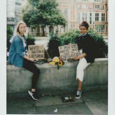 Two people sit on a low wall, holding signs which say "White inaction is lazy violence" and the other says "If 'all' lives matter why are you outraged".