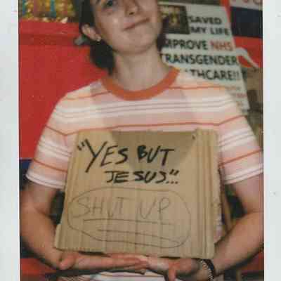 A person wearing a cap holds a cardboard sign with handwritten messages, smiling gently at the camera.