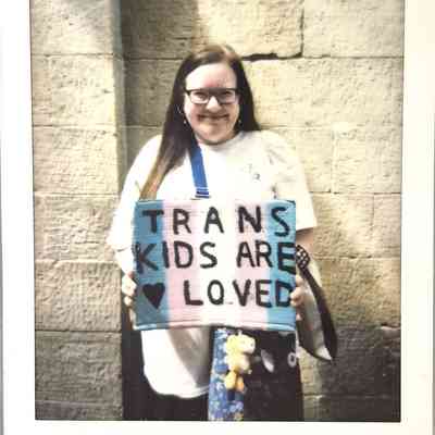 A smiling person holds a colorful sign reading "TRANS KIDS ARE LOVED" against a stone wall