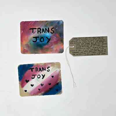 Two colourful fabric patches, one in the trans flag colours, both with "TRANS JOY" written on them.  With a brown, handwritten luggage tag.