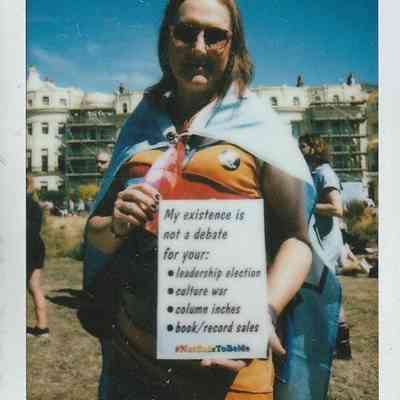 A person wearing sunglasses and a cape holds a sign which says "My Existence Is Not A Debate For Your -leadership election -culture war -column inches -book/record sales #NotSafeToBeMe'.