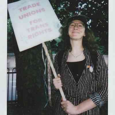 A person is standing outside holding a sign that reads "Trade Unions for Trans Rights" and smiling.