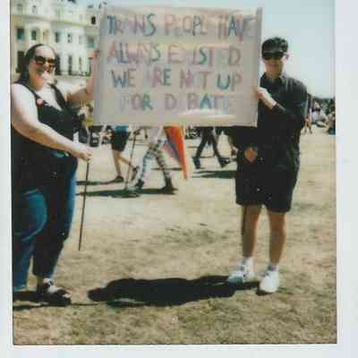 Two individuals hold a sign that says "Trans people have always existed. We are not up for debate".