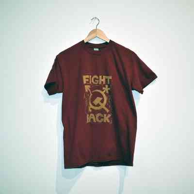 A red t-shirt printed in gold with the trans symbol stylised as a hammer and sickle and the text "FIGHT BACK". On a wooden hanger.