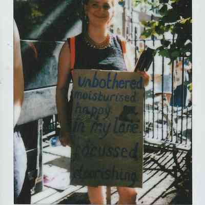 A person stands outside holding a placard which says "Unbothered, moisturised, happy, in my lane, focused, flourishing".