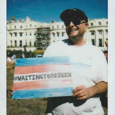 A person holds a colorful sign with "#WAITINGTOBESEEN" outdoors by a large building.