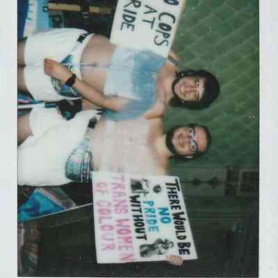 Two individuals hold protest signs advocating for Pride inclusion and celebrating trans women of color.