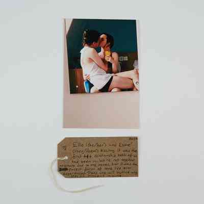 Photo of two dark haired people, one sitting the the others lap, kissing. With a brown, handwritten luggage tag.