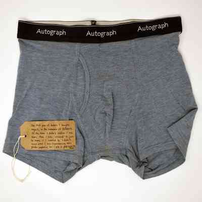 A pair of grey boxer shorts with a black waistband displaying the repeated text "Autograph". With a brown handwritten luggage tag.