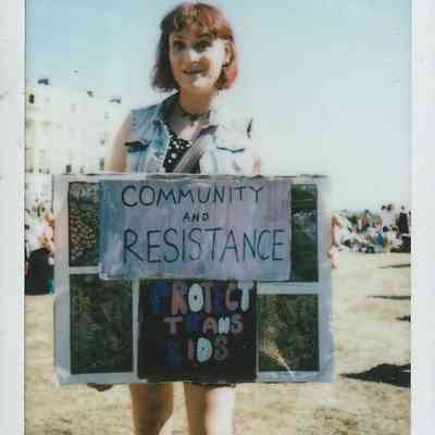 A person stands holding a sign which says "COMMUNITY AND RESISTANCE PROTECT TRANS KIDS".
