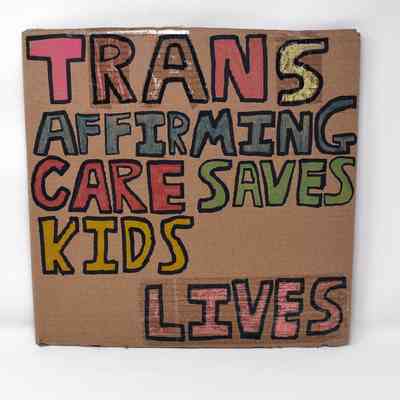 A placard made from cardboard which has 'Trans Affirming Care Saves Lives' in colourful block text.