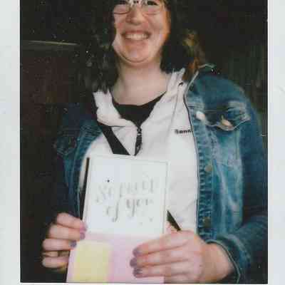 A person smiling holds a card reading "So Proud of You," wearing a denim jacket in Leeds '