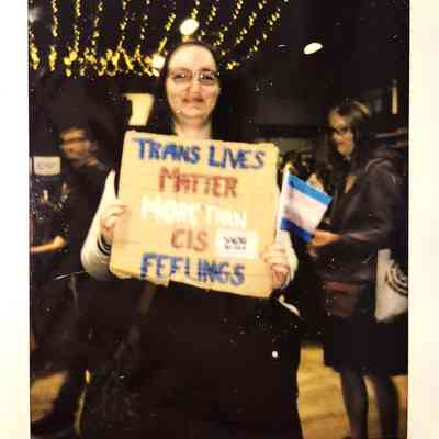 A person holds a sign saying "Trans Lives Matter More Than Cis Feelings" at an indoor gathering.