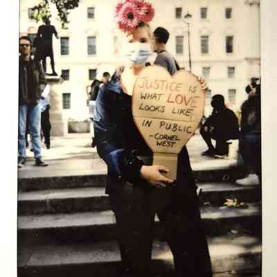 A person wearing a mask and flower headdress holds a heart-shaped sign.