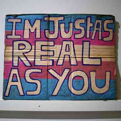 A hand painted cardboard placard in the trans flag colours and the text "IM JUST AS REAL AS YOU"