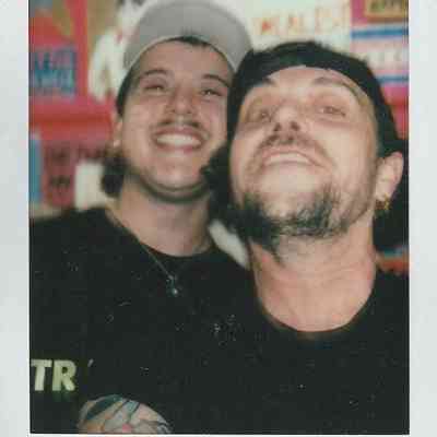 Two people are smiling at the camera in a colorful room with various posters in the background.