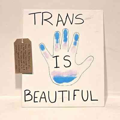A placard which is white, has an outline of a hand drawn and inside is painted in trans flag colours - blue, pink and white. It has 'Trans is Beautiful' written on it. Lay on the placard is a brown tag with writing on it.