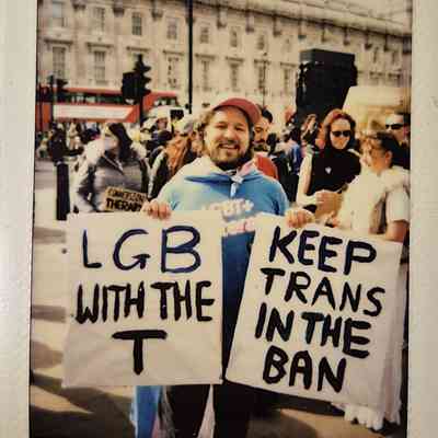 A person at a protest holds signs which say "LGB WITH THE T" and "KEEP TRANS IN THE BAN".