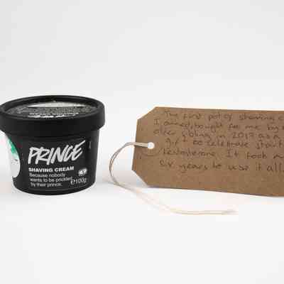 A black tub of 'Prince' Shaving Cream from Lush. There is a brown tag with writing on it next to it.