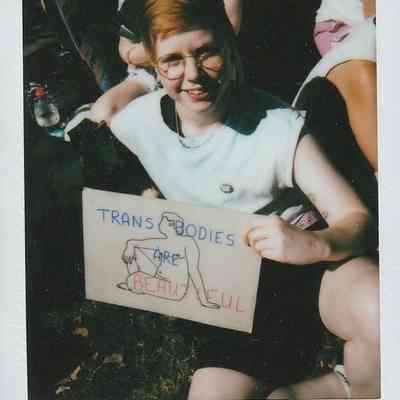 A person with glasses holds a sign saying "Trans Bodies Are Beautiful" while sitting on the grass outdoors.